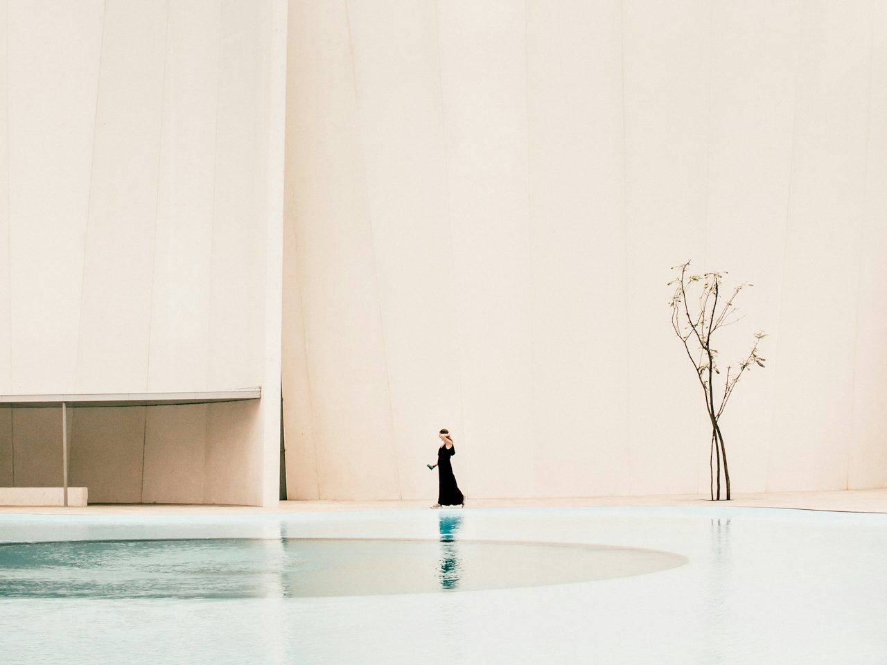 Femme en noir près d'une piscine, avec un arbre sec en arrière-plan sur un fond clair.